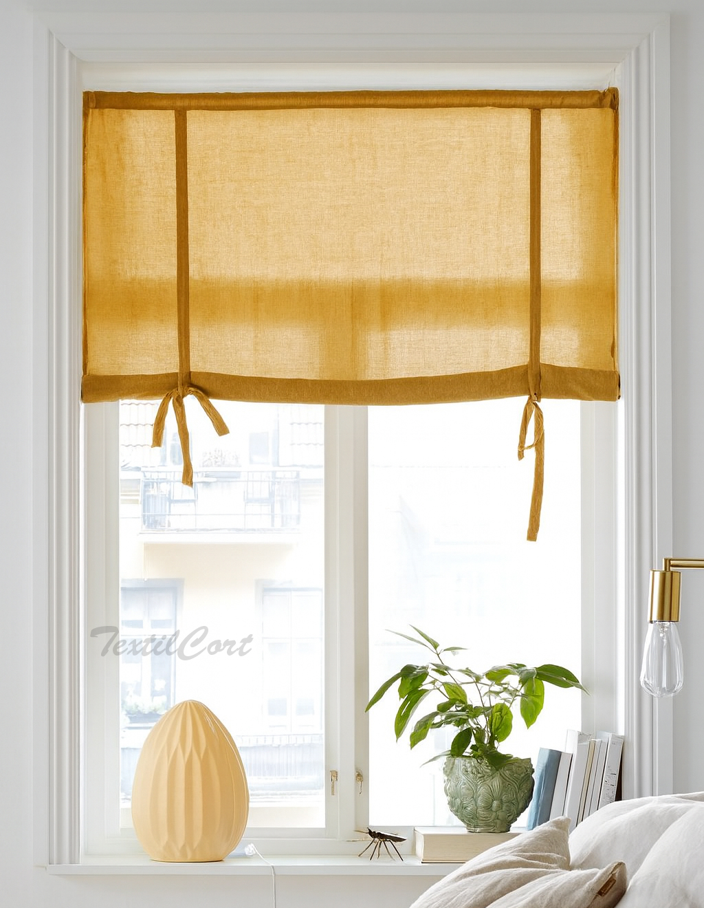 Estor con lazos decorativos en tejido mostaza translúcido sobre ventana blanca en dormitorio luminoso de estilo nórdico