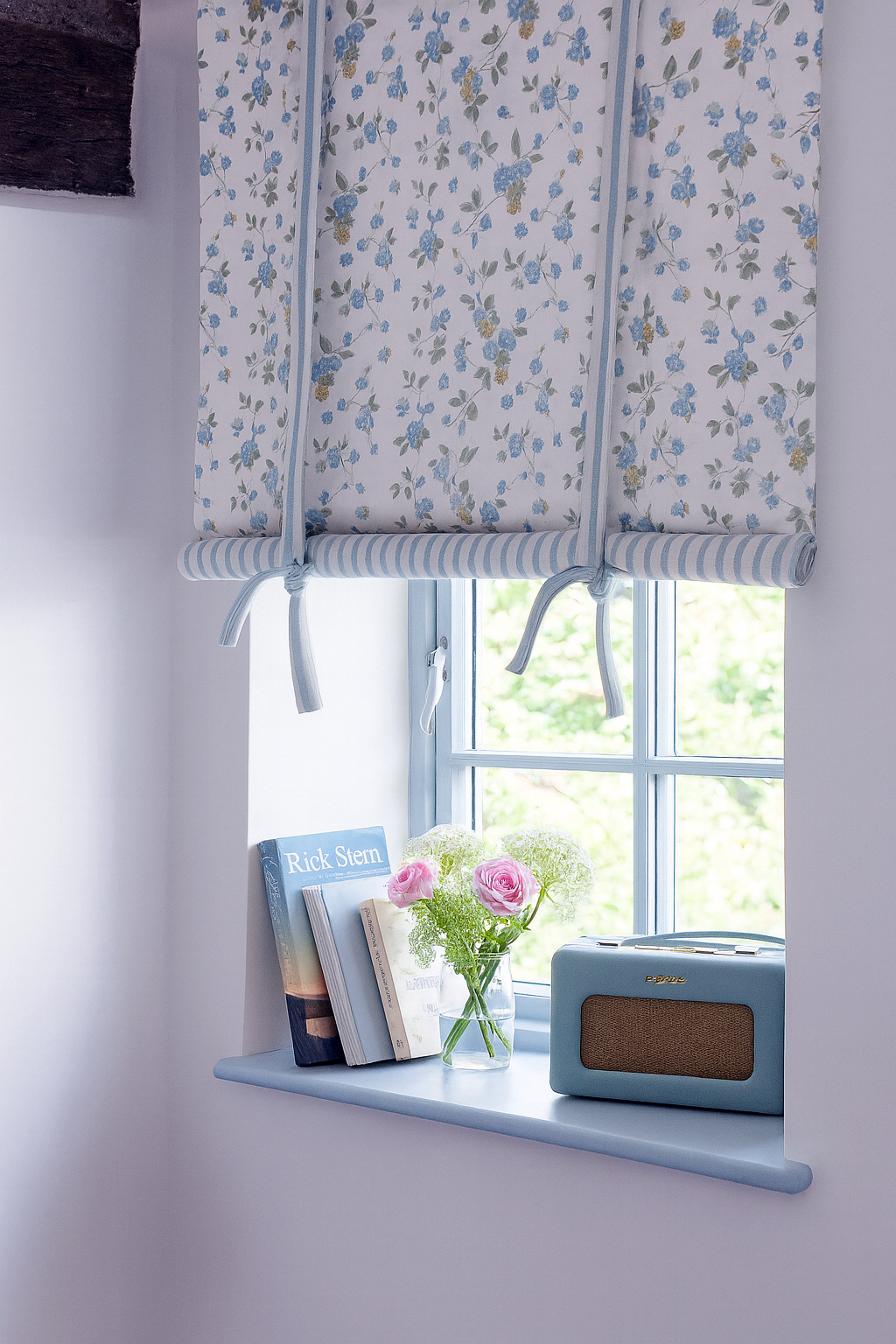 Estor con estampado floral azul sobre fondo blanco con lazos decorativos, en ventana pequeña con libros y flores